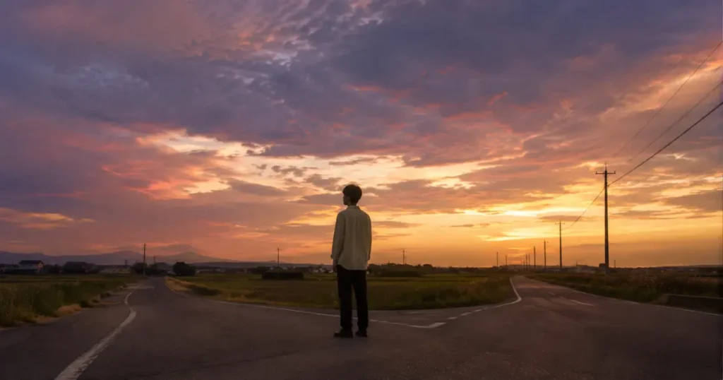 夕暮れの分かれ道に立ち、空を見上げる若者——自分の立場と時間を見つめ直す