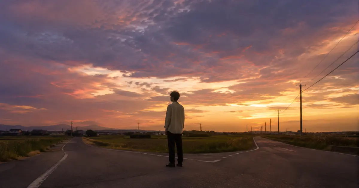 夕暮れの分かれ道に立ち、空を見上げる若者——自分の立場と時間を見つめ直す